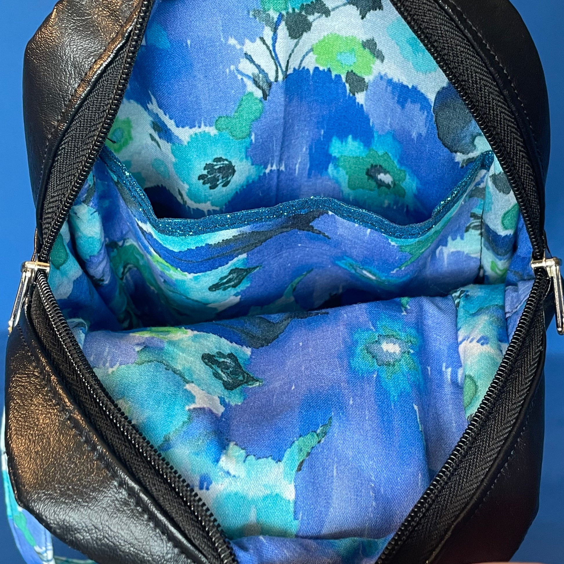 Beautiful interior view showing the blue and green watercolor lining fabric. Black reclaimed vinyl from scraps of car interior. Showing interior slip pocket with a surprise of blue sparkly pocket trim. Highlights the double zipper opening and spacious compartment.